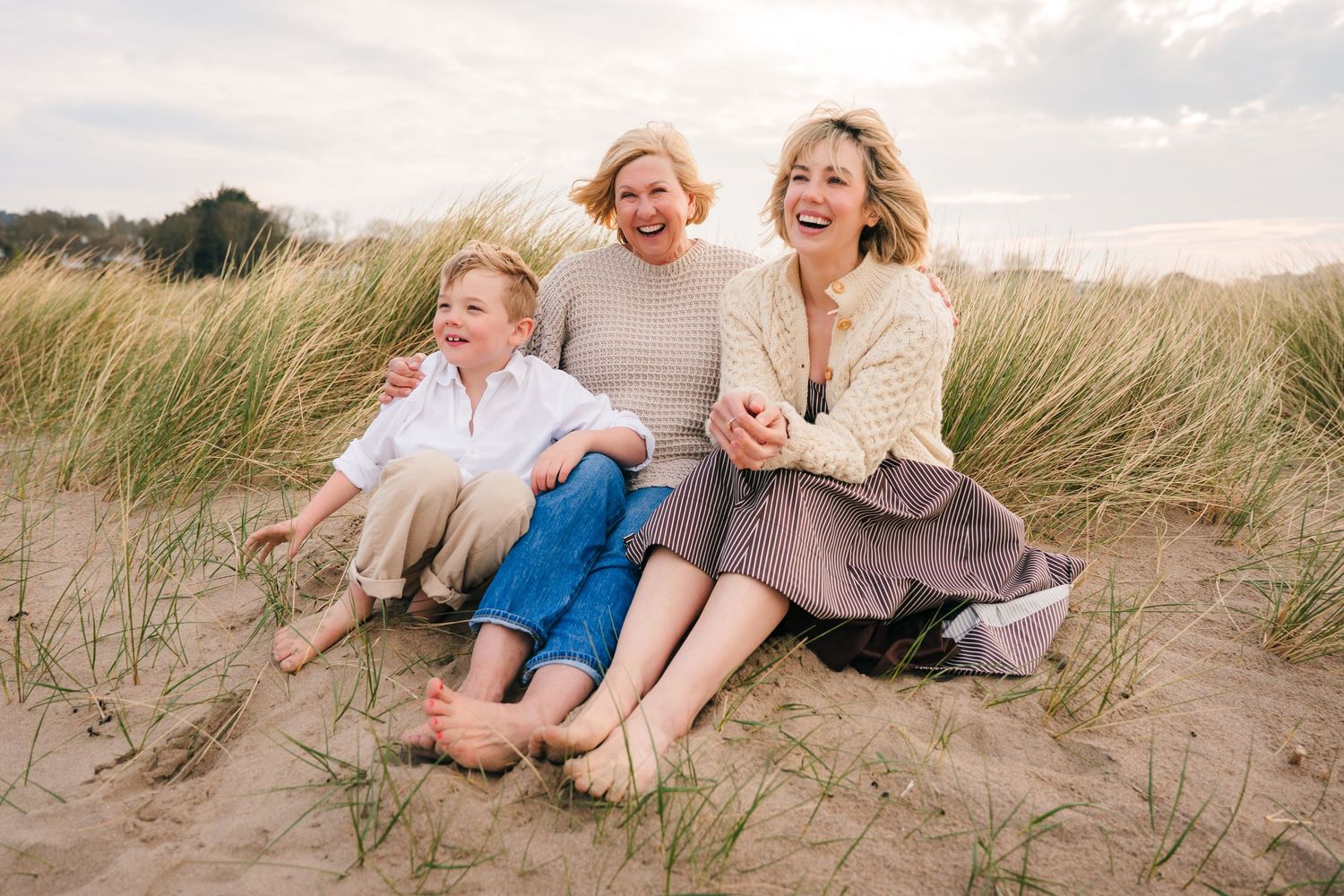 A family enjoys a candid moment together while sitting in the grassy sand dunes at sunset. Family PHOTOGRAPHER Dublin