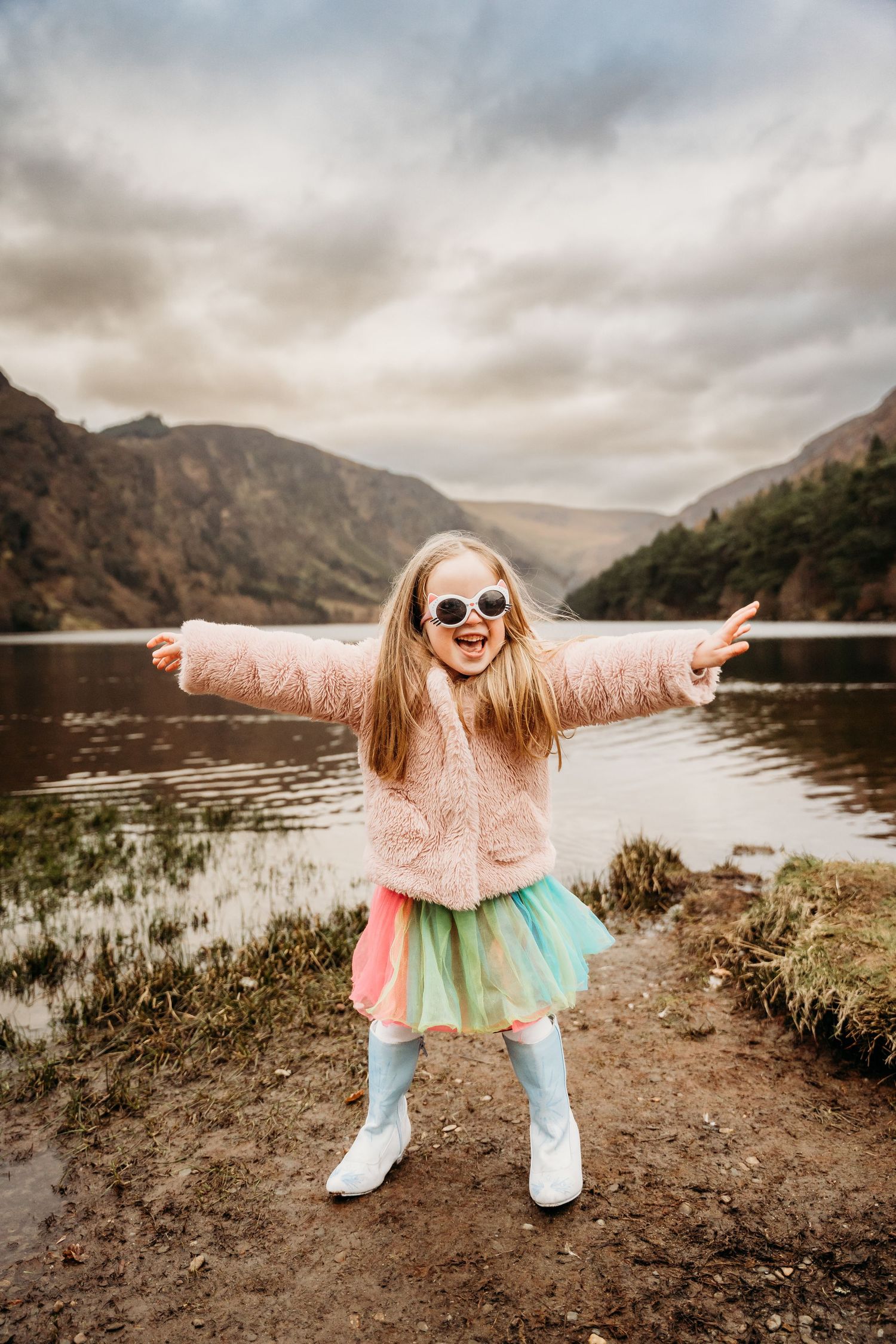 Joyful child in pink sweater and turquoise tutu dances by a scenic mountain lake on a cloudy day. Family PHOTOGRAPHER Dublin