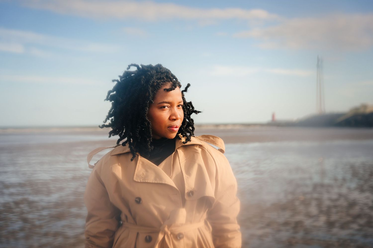 Person in beige coat standing thoughtfully on sandy beach with cloudy sky. Dublin professional headshots photographer