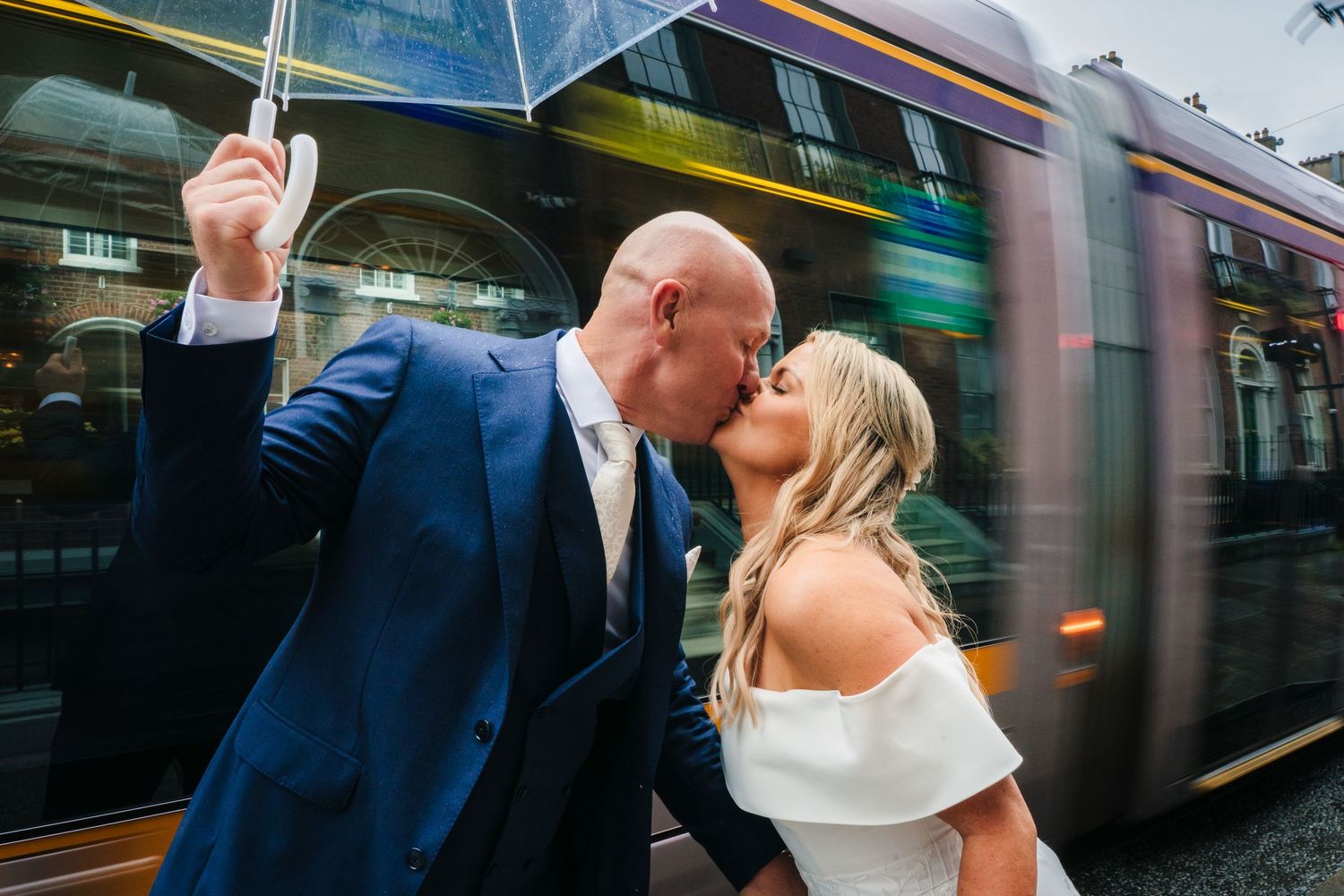 A couple shares a romantic kiss near a moving transit train in an urban setting. The Dean Dublin Wedding Photographer