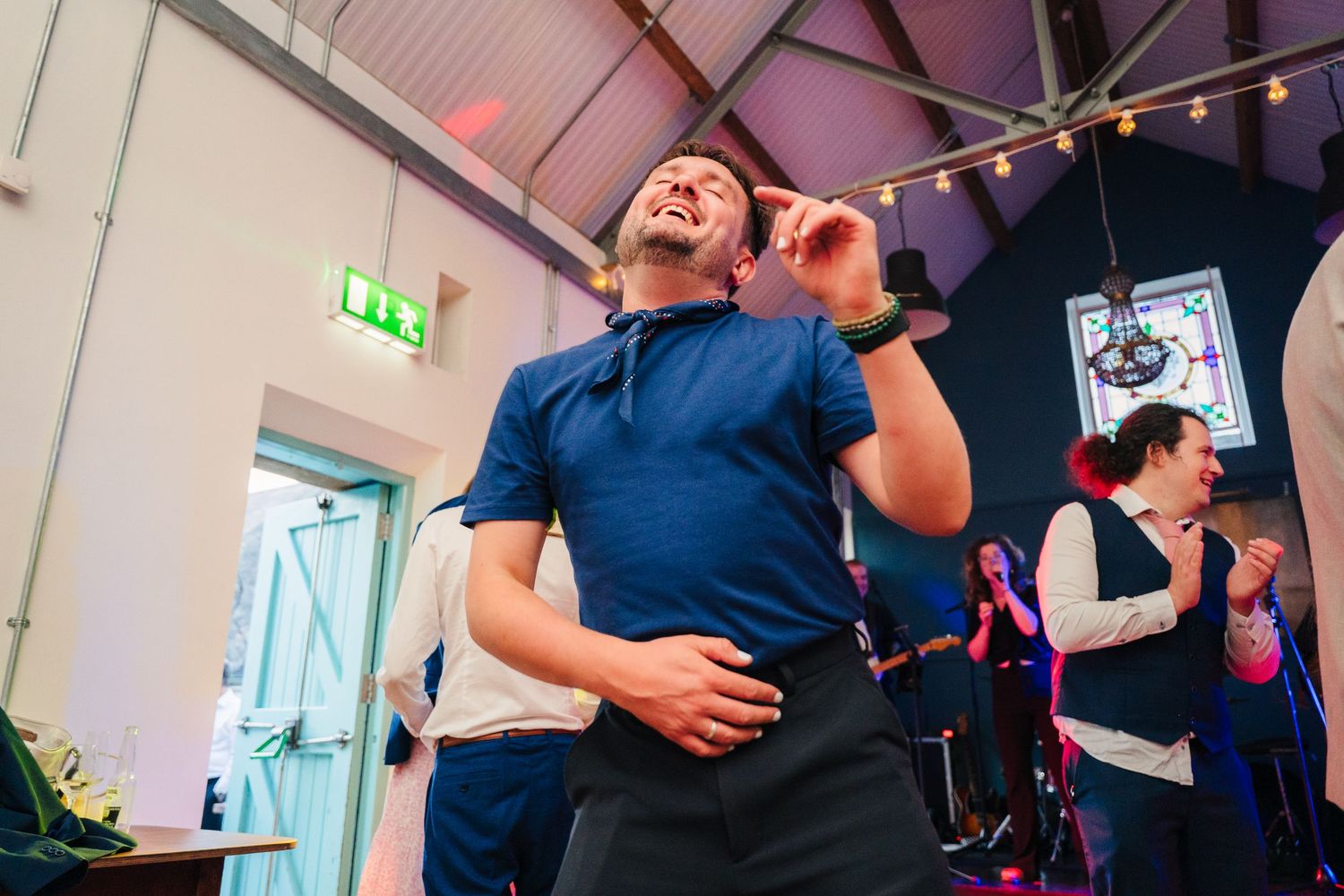 An enthusiastic dancer moving energetically on a wooden dance floor under string lights at an indoor event. . Village Barn Tyrrellspass wedd