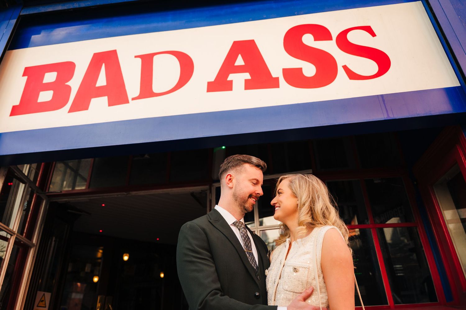 Couple posing romantically under illuminated BAD ASS neon sign at night. City Hall Dublin wedding photogrAPher