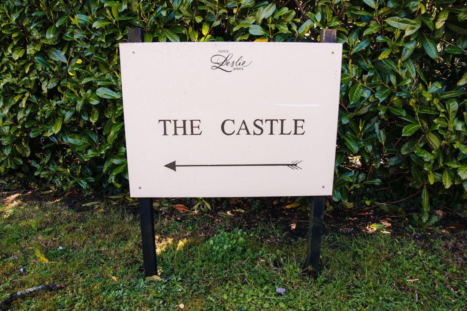 A white directional sign for 'The Castle' mounted on a black post stands in front of lush green hedges. Castle Leslie Wedding Photographer D