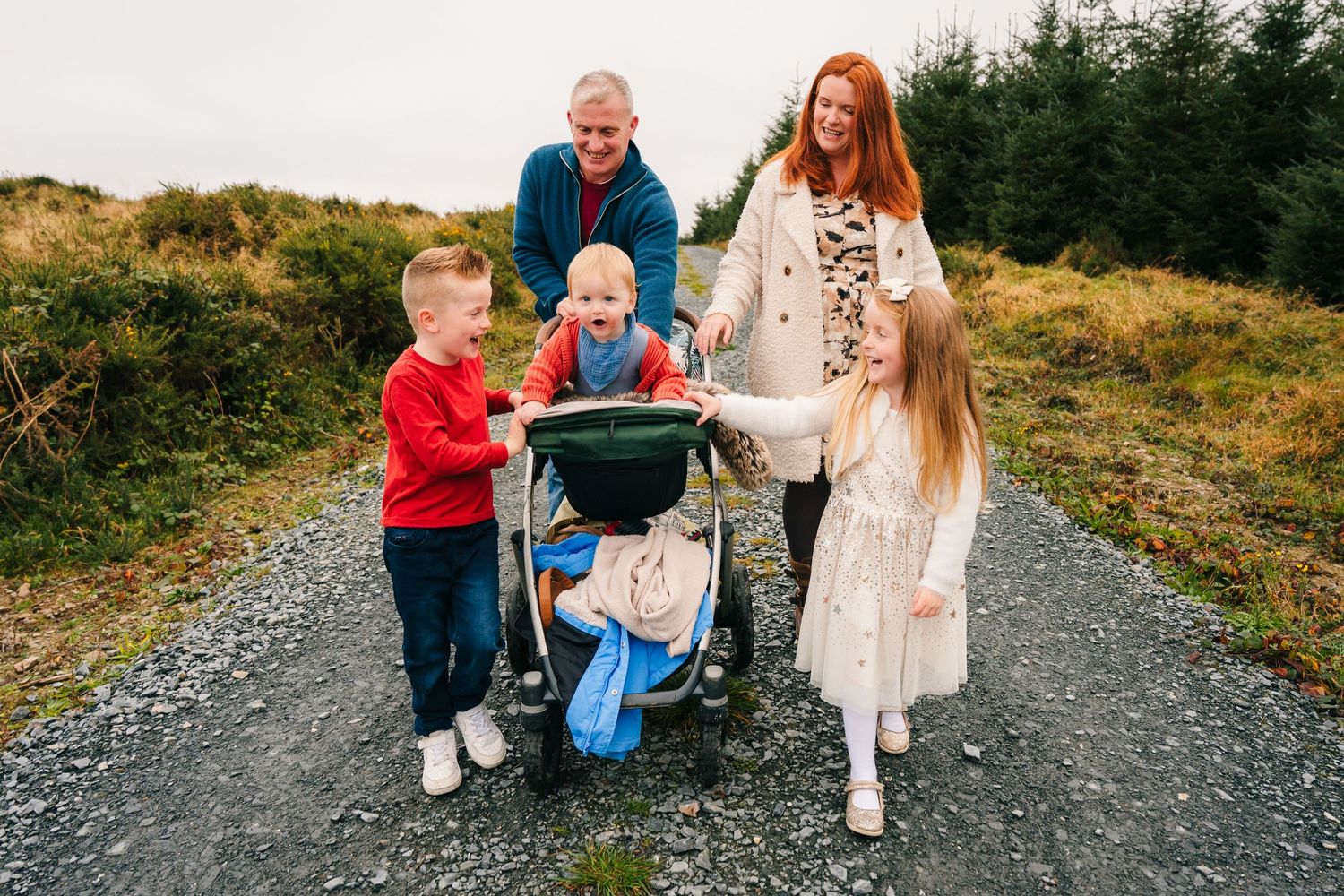 Family Photographer Dublin Ireland A family enjoys a playful moment together on a gravel path surrounded by evergreen trees.