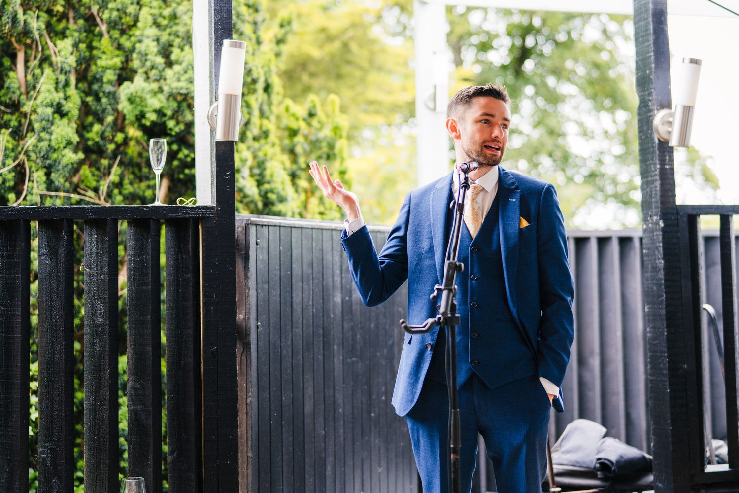 Poulaphouca Wedding photographer wicklow  Speaker in a navy blue suit addresses wedding guests from an outdoor deck.