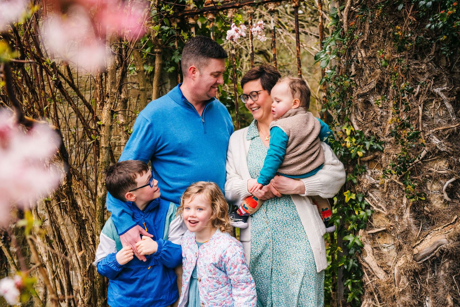 Family of five poses together outdoors in spring with pink blossoms and natural foliage backdrop. Family photographer Dublin Ireland