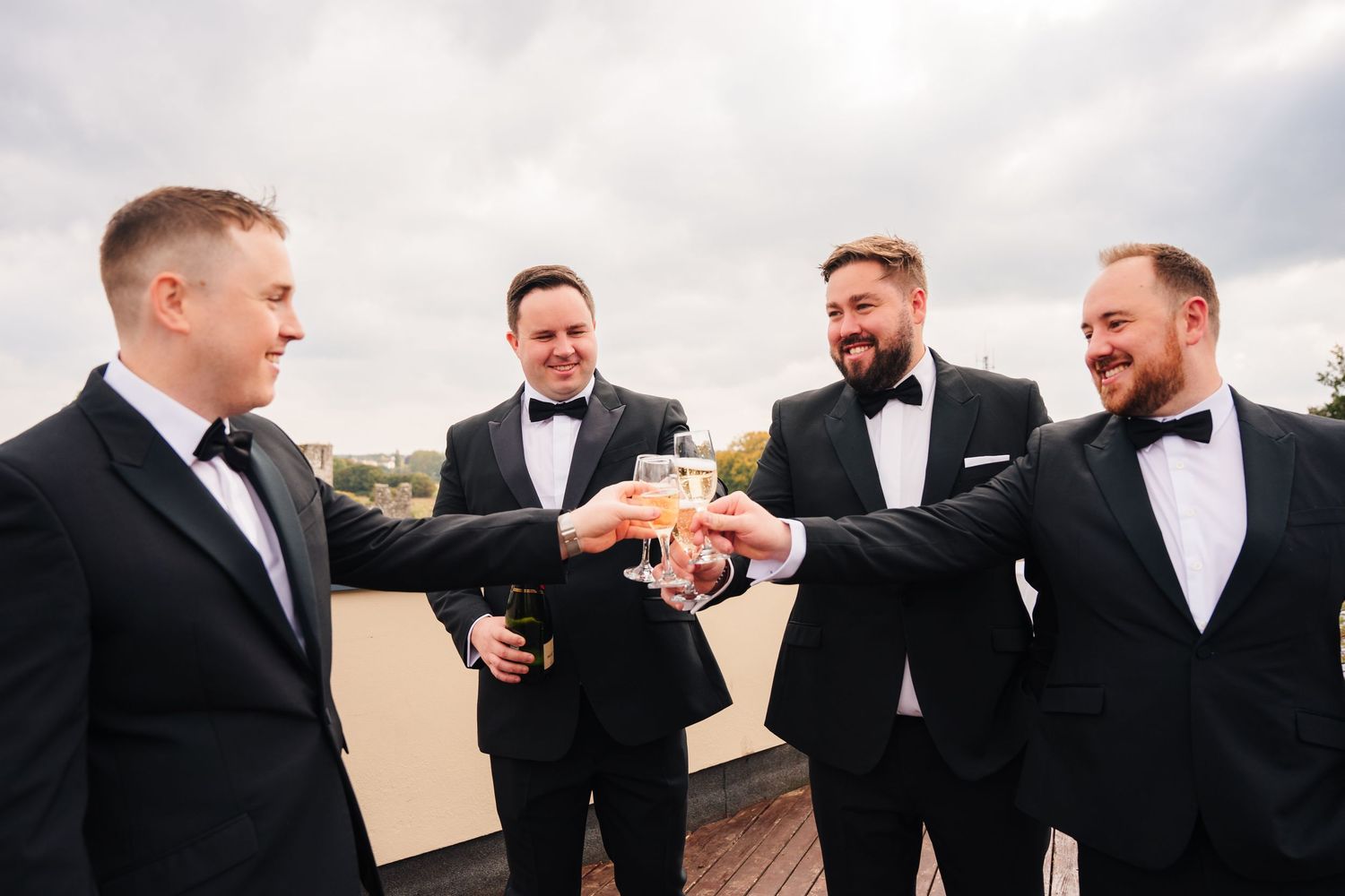 A group of groomsmen in black tuxedos and bowties share a laugh while gathered together outdoors. Wedding Photography Trim Castle Ireland