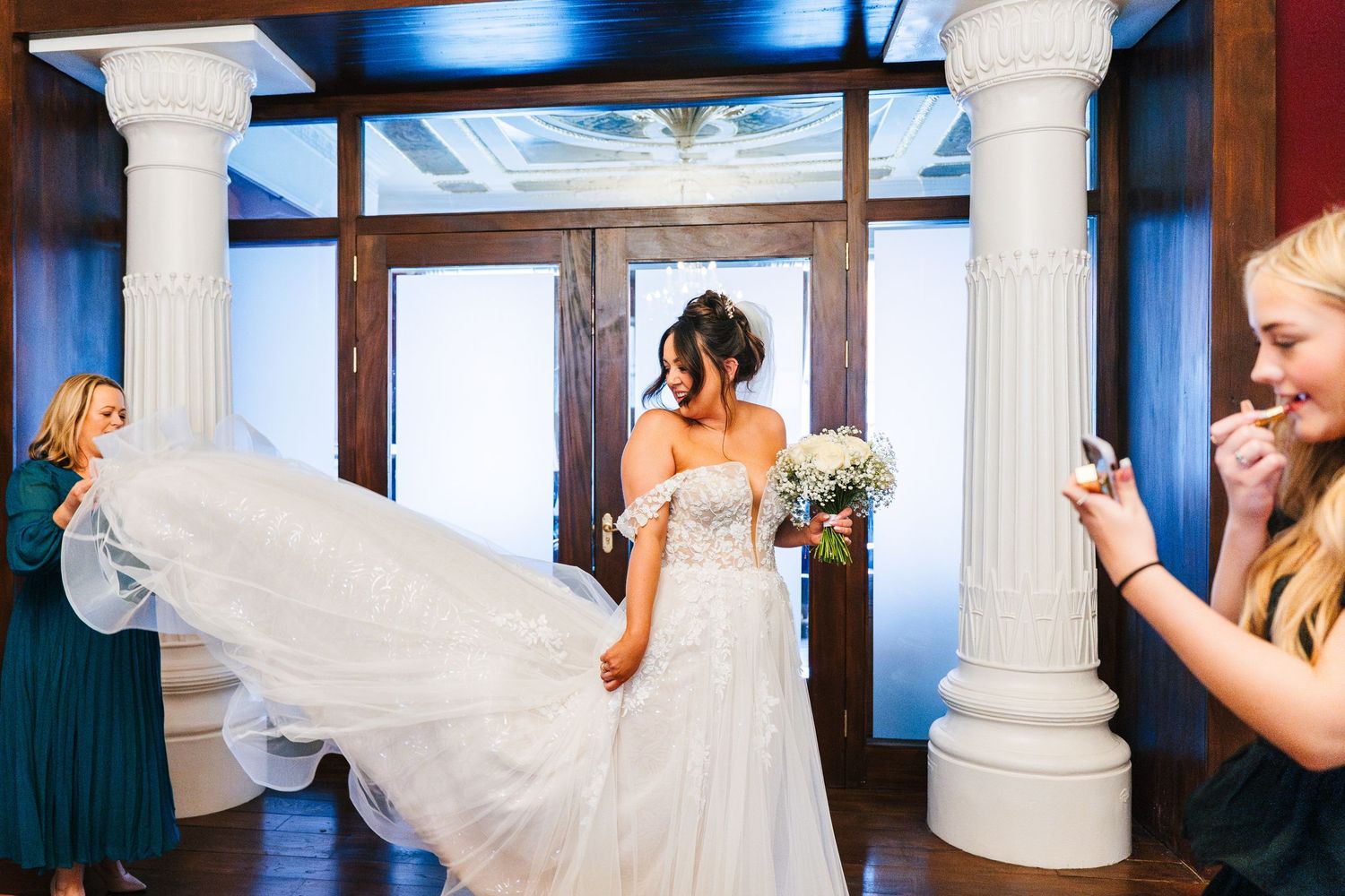 A bride in a flowing white wedding dress spins joyfully between elegant white pillars as others look on.  Wedding PHOTOGRAPHER Dublin