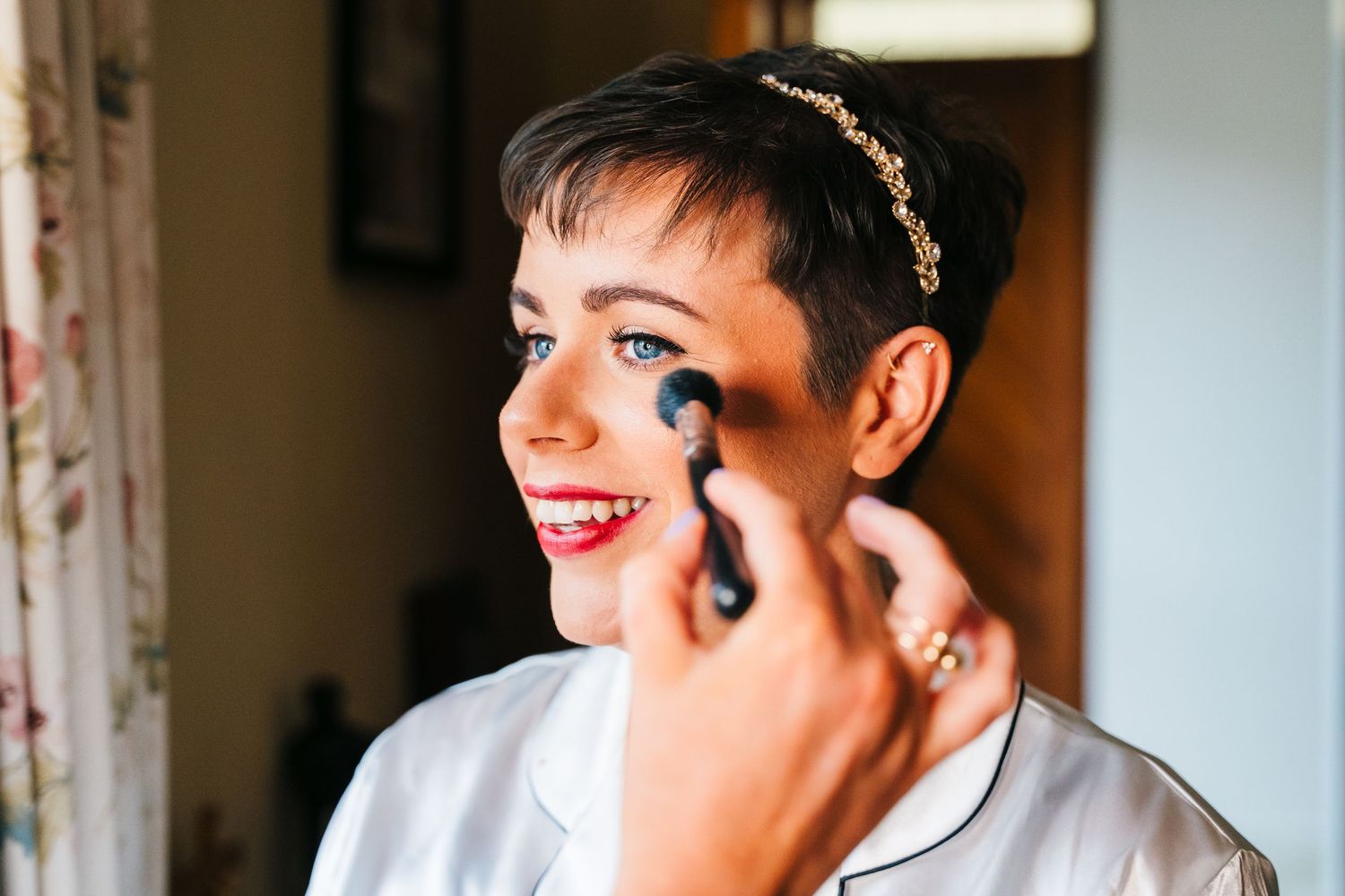 Makeup artist applies cosmetics to a smiling person's face before a special event in a warmly lit room. Millhouse slane wedding photographer