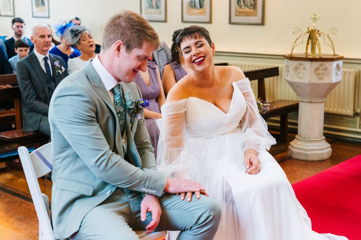 Happy couple shares joyful laughter during wedding ceremony in traditional church setting with baptismal font visible.Millhouse slane weddin