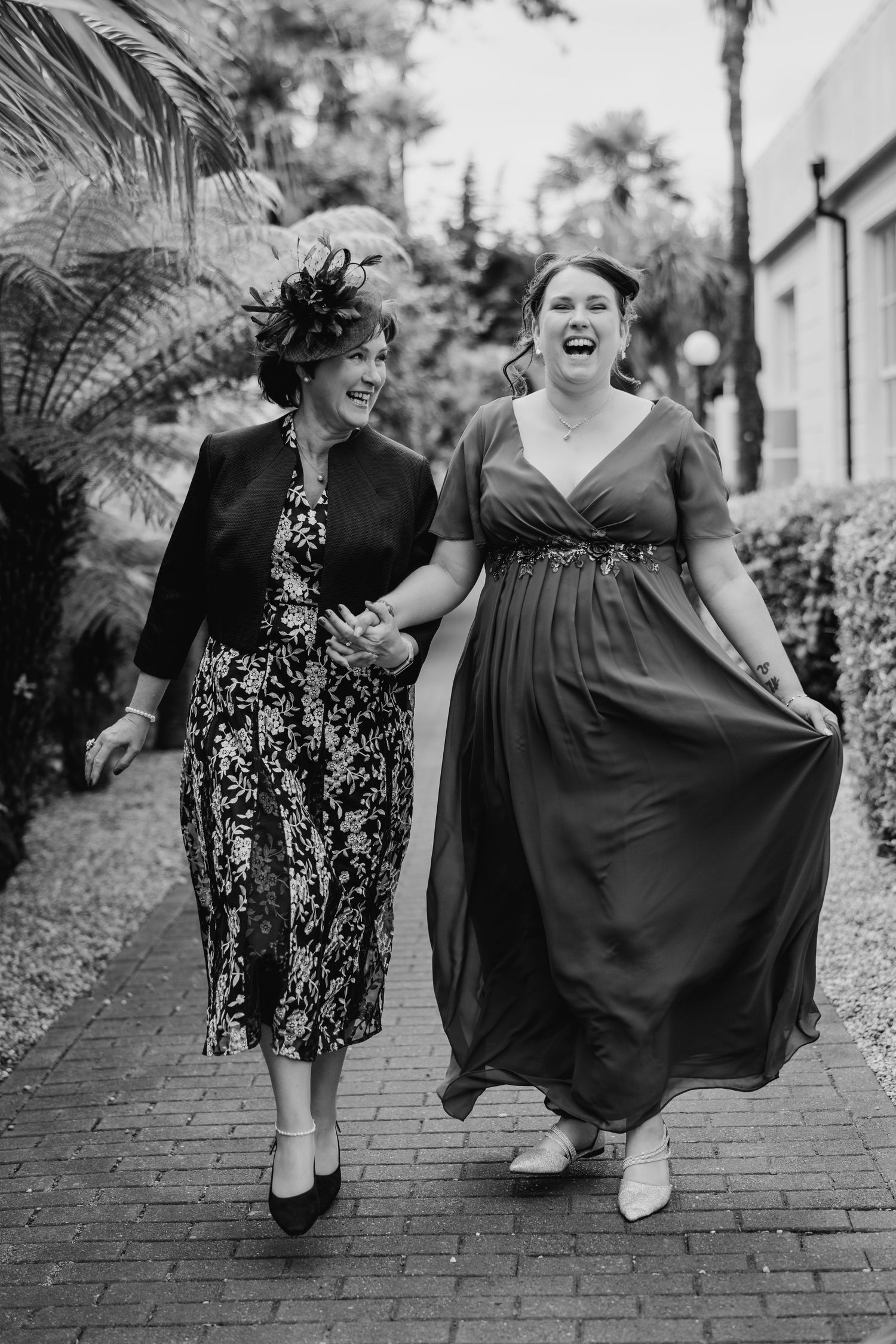 Two women laughing and walking together on a brick path, one in a floral dress and one in an elegant evening gown. Portmarnock Hotel Wedding
