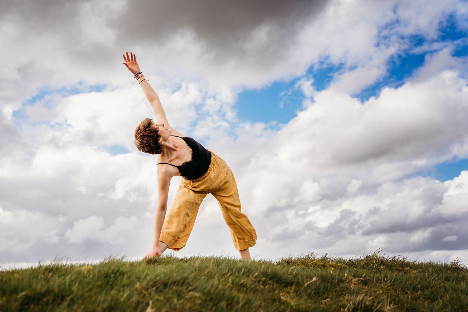 Person in yellow pants performs a yoga pose on a grassy hilltop against a dramatic cloudy sky. Portrait PHOTOGRAPHER Dublin