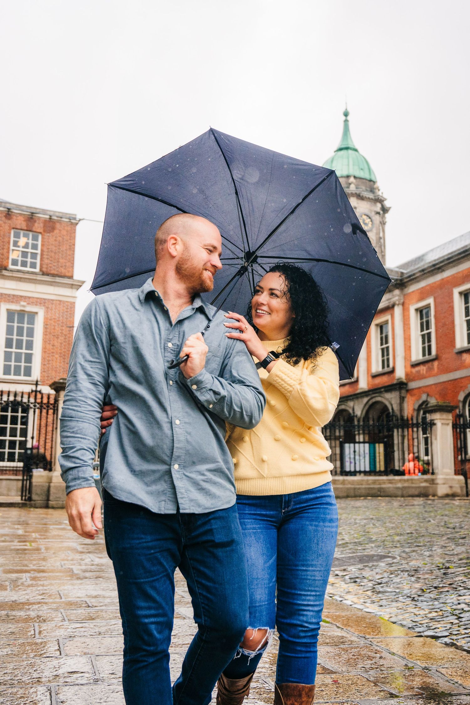 A couple shares a romantic moment under a black umbrella against red brick buildings on a rainy day. Proposal Dublin Photographer