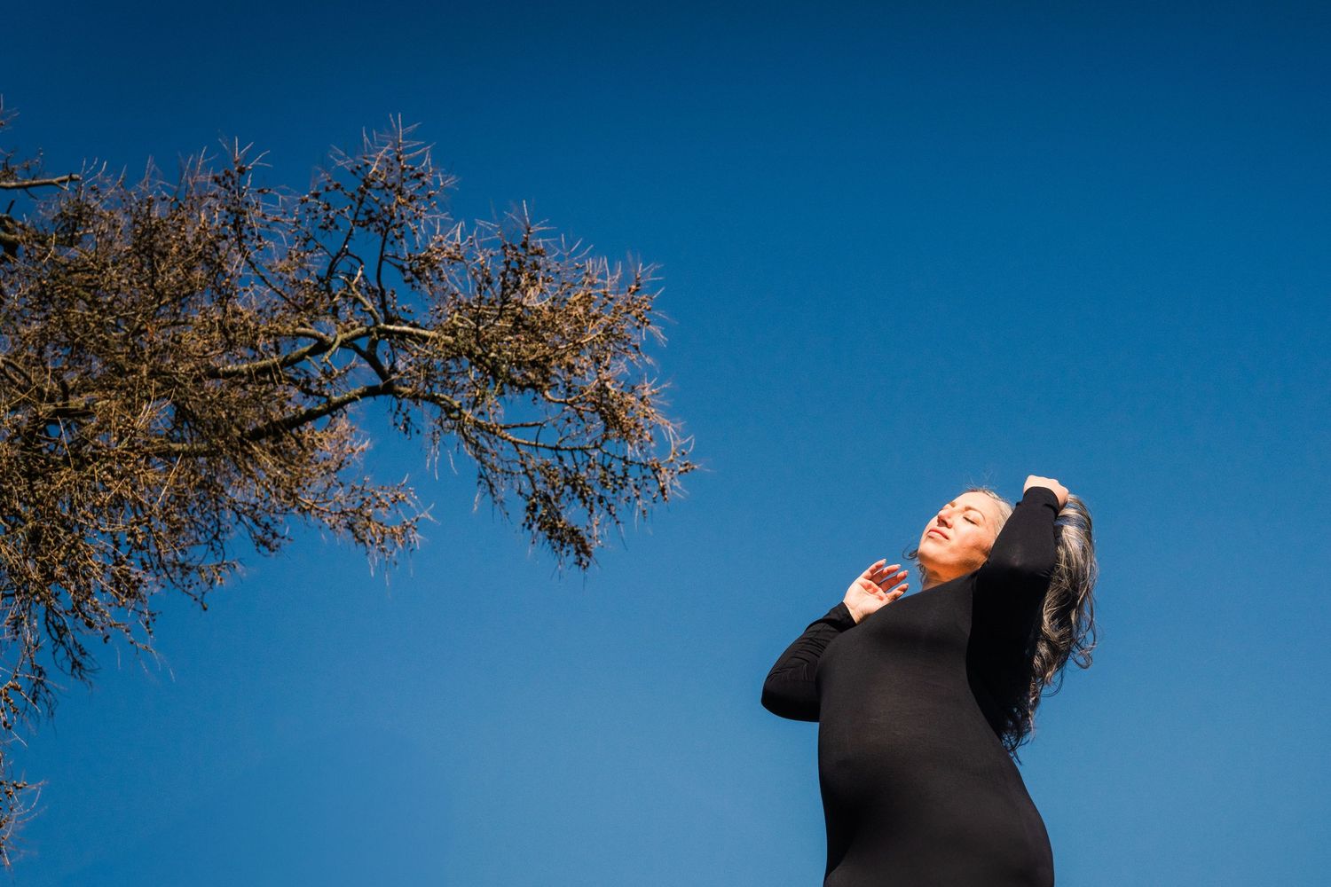 Person in black stretches joyfully against bright blue sky with autumn tree branch overhead. Family PHOTOGRAPHER Dublin