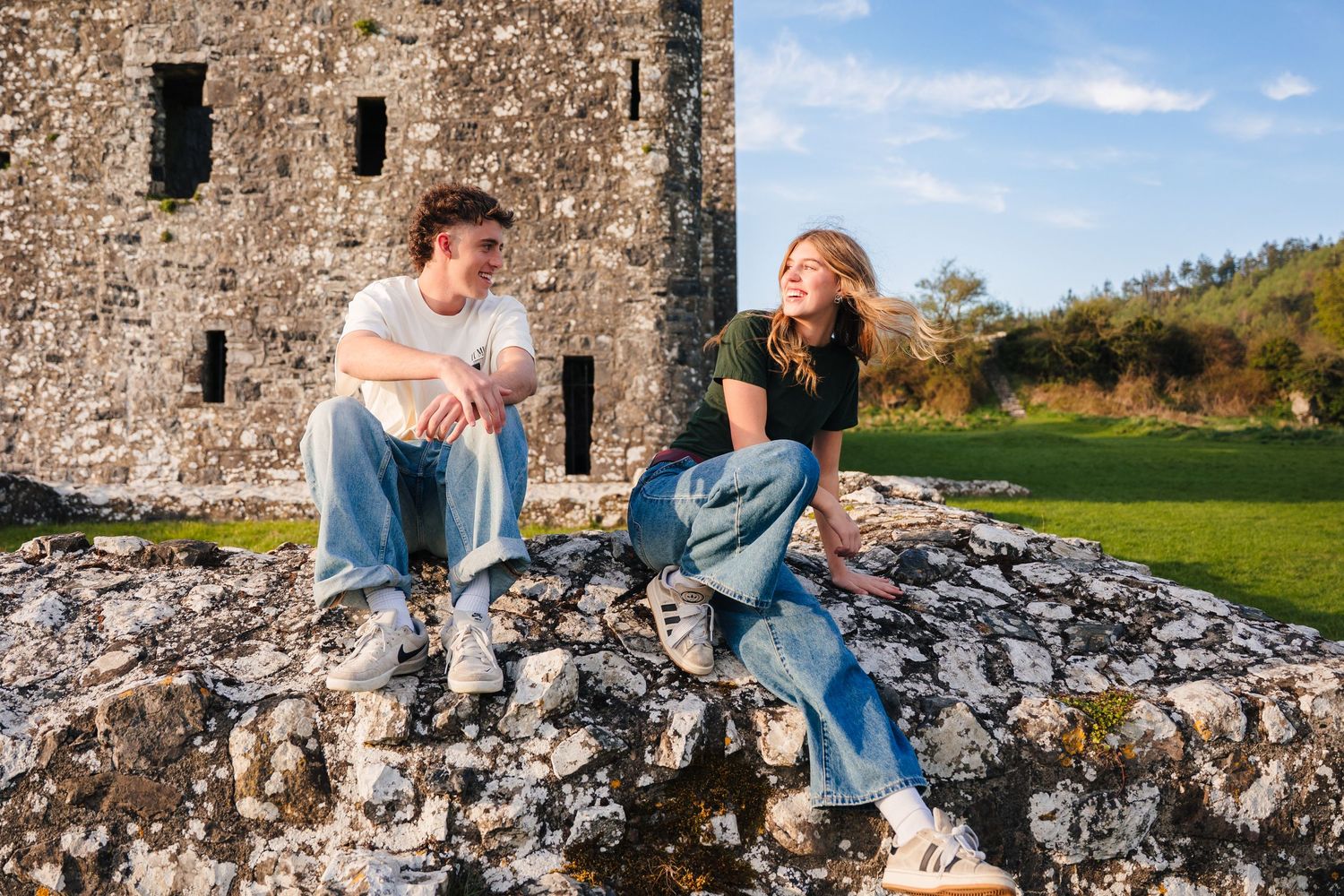 Two people sit relaxing on stone ruins near an old castle tower on a sunny day in the countryside. Photographer Ireland dublin