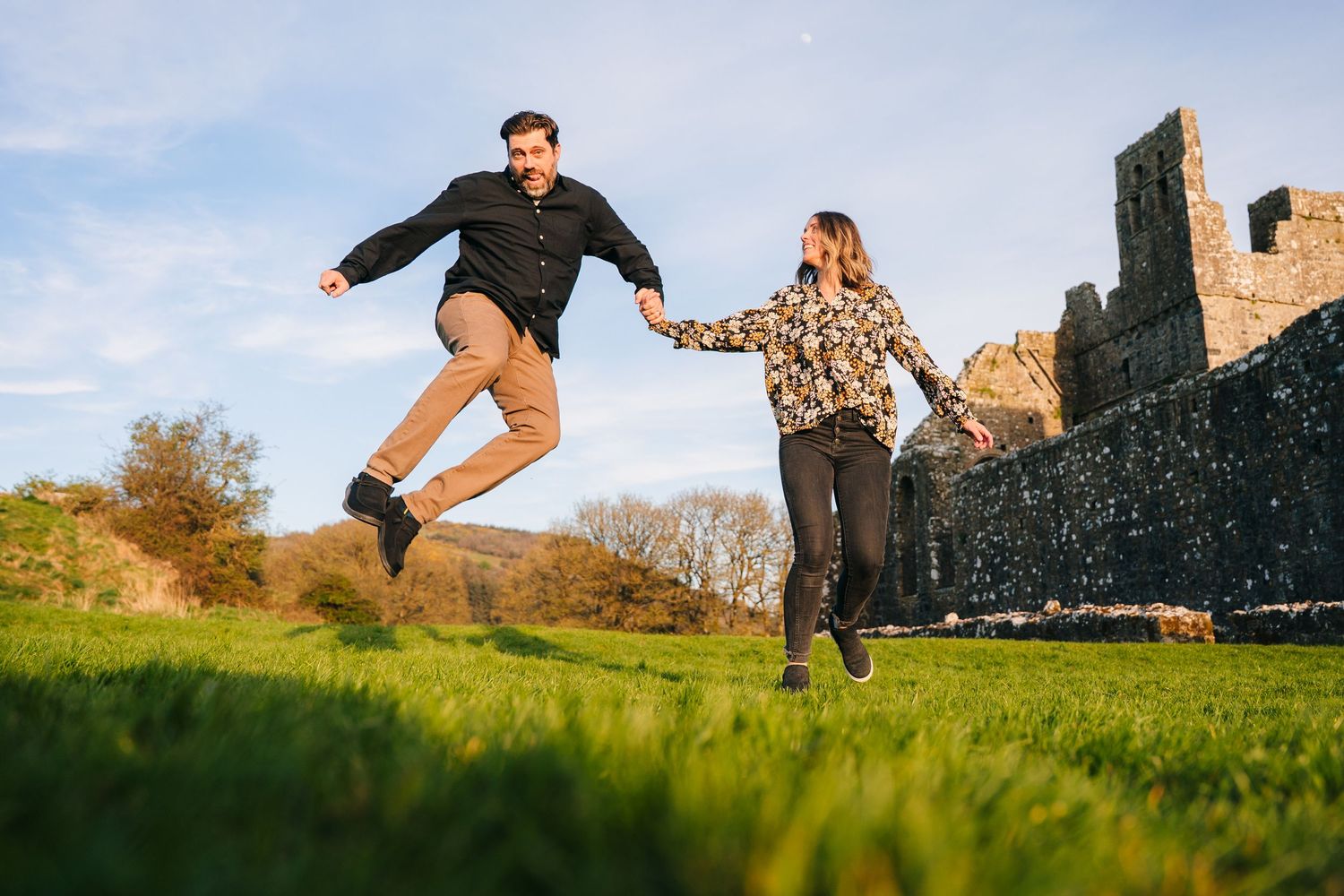 Two people jumping joyfully on a green hill with ancient castle ruins in the background on a sunny day. Photographer Ireland dublin