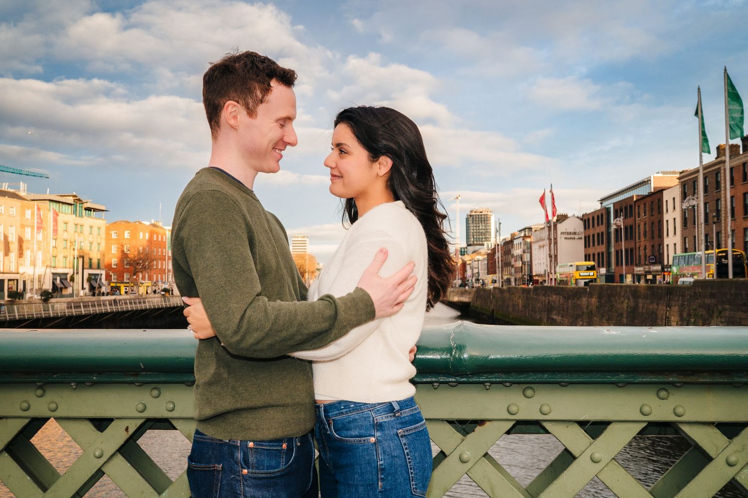 Couple in casual sweaters embracing on bridge with colorful Dublin buildings in background. Surprise proposal dublin photography