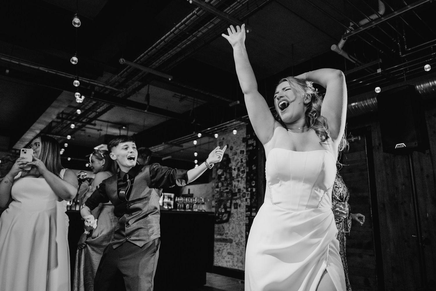 Black and white photo of wedding guests dancing joyfully with arms raised at an indoor reception venue. Wedding Photographer Dublin