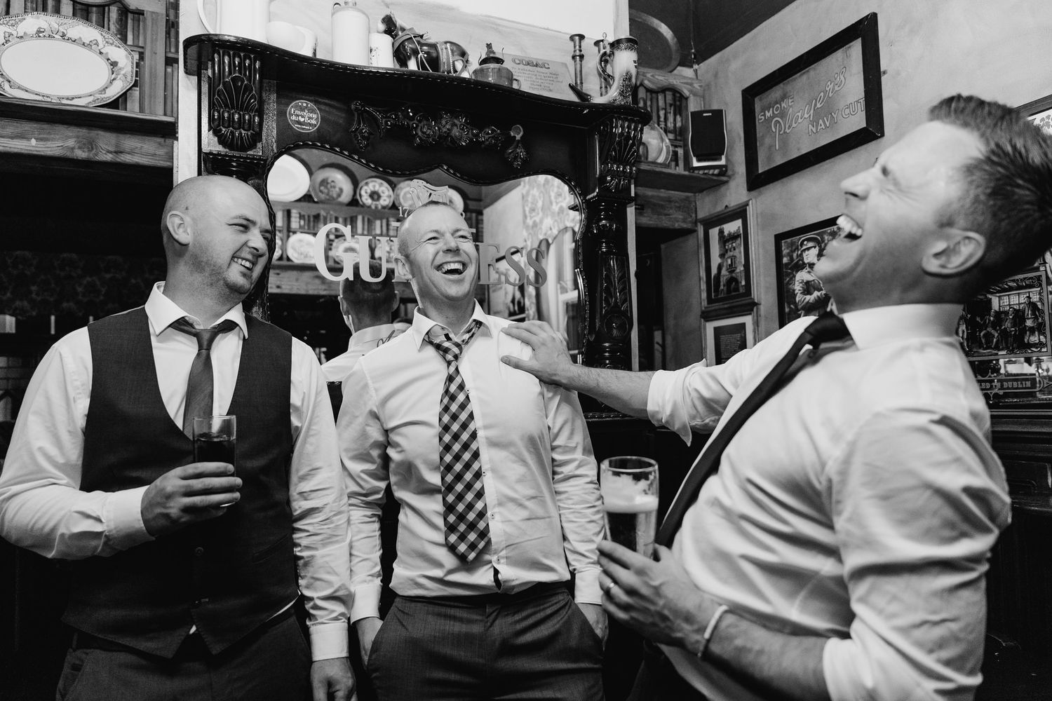 Three men laugh together at a pub while holding drinks in a black and white candid photograph. Wedding PHOTOGRAPHER Dublin