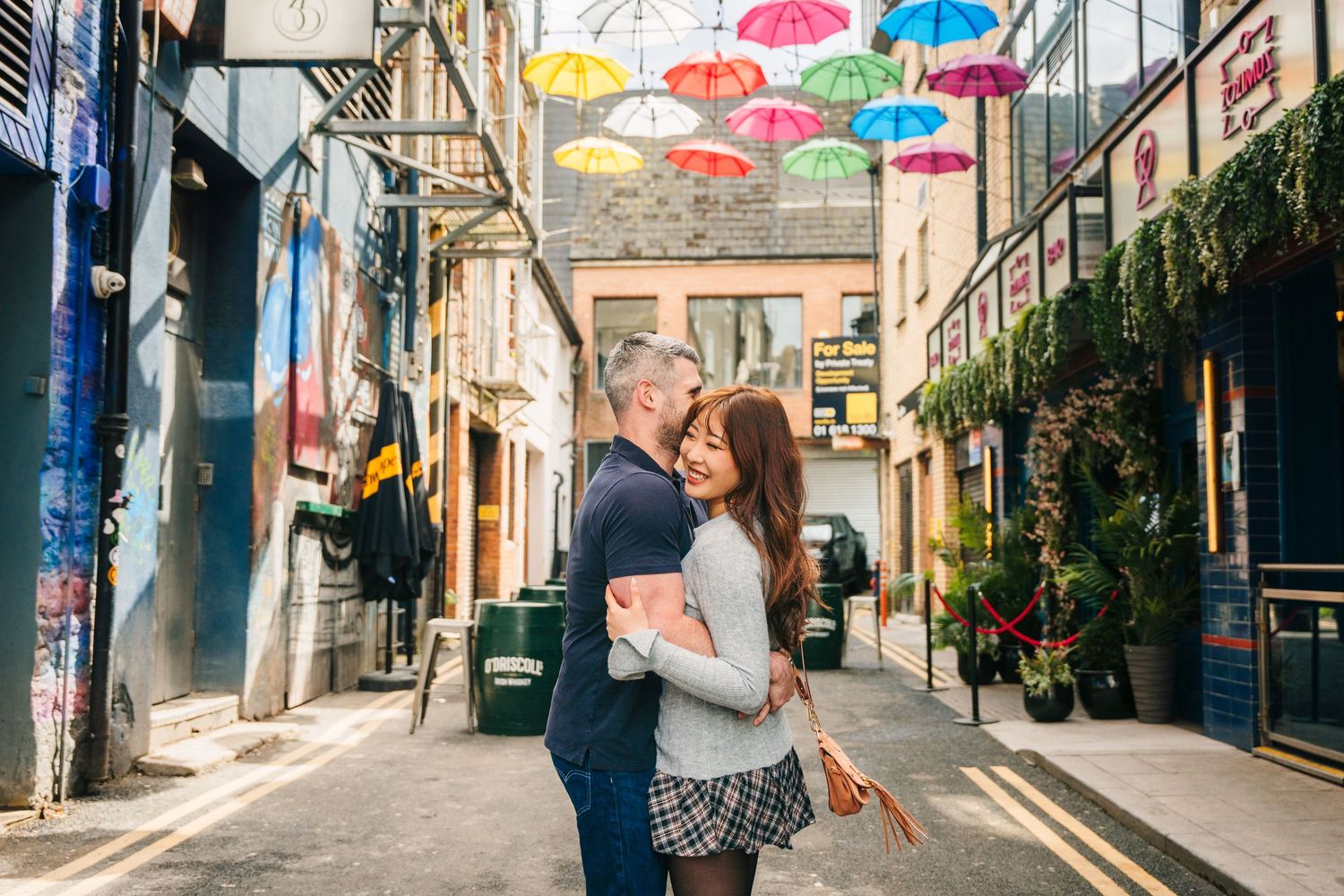 A romantic moment under colorful floating umbrellas in a charming alleyway decorated with hanging plants and flowers. PHOTOGRAPHER Dublin