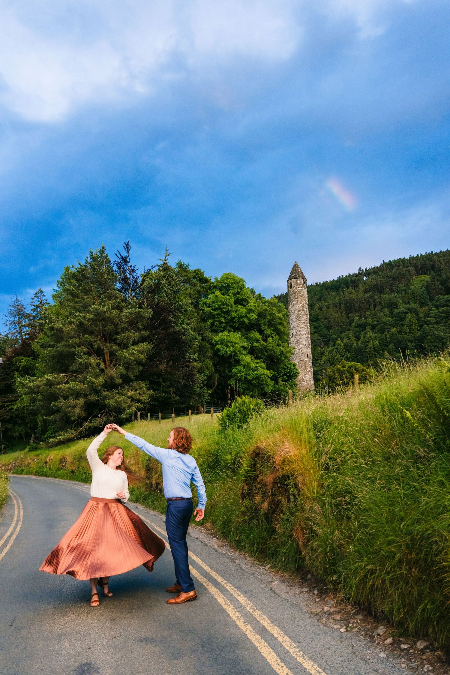 Two people dance joyfully on a winding road near an ancient stone tower in Ireland's lush countryside under blue skies. PHOTOGRAPHER Dublin