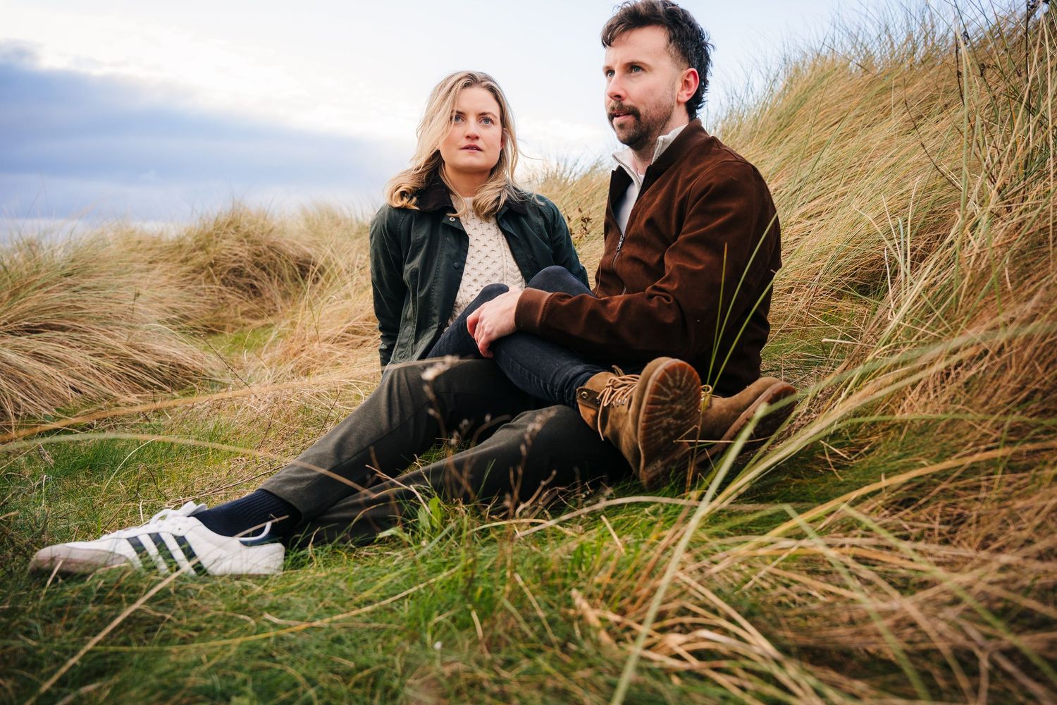 Two people sit together in tall grass on a coastal sand dune on an overcast day. PHOTOGRAPHER Dublin ireland