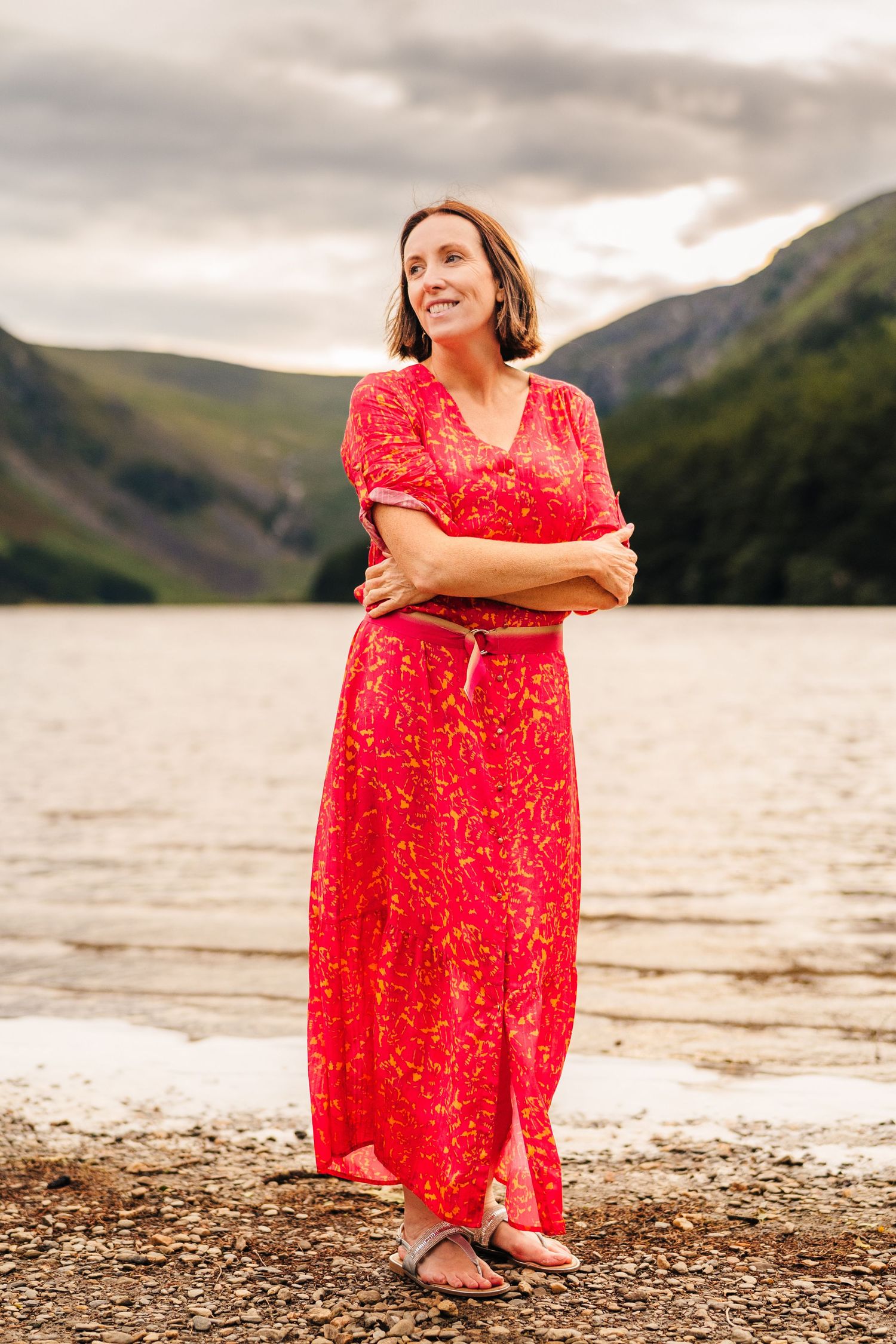 Person in flowing red dress poses along rocky lakeshore with misty mountains in background at sunset. portrait PHOTOGRAPHER Dublin