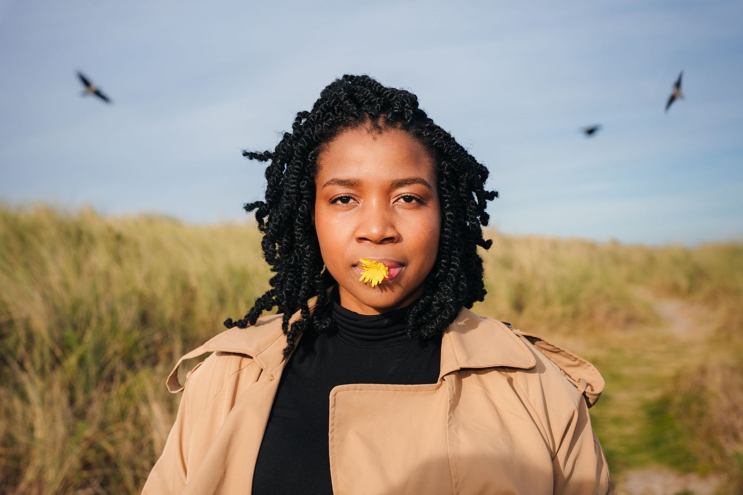 Person in a tan coat poses in a meadow with birds flying overhead in the blue sky. PHOTOGRAPHER Dublin