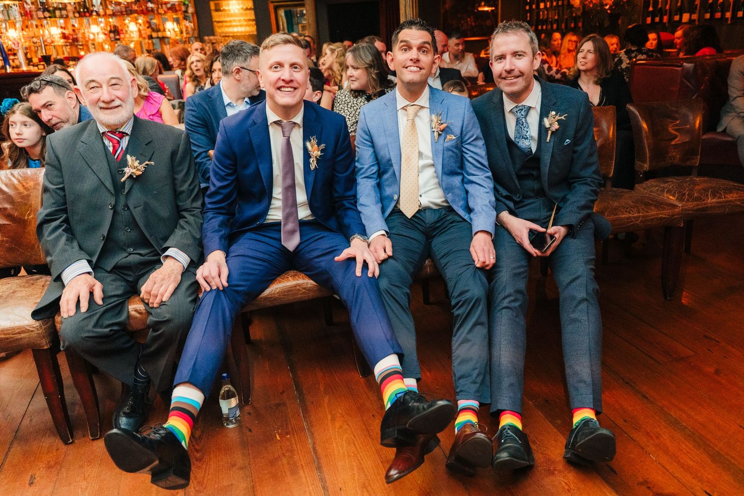 Well-dressed wedding guests in blue and gray suits sitting together on a wooden bench at reception. Marco Pierre Dublin Wedding Photographer