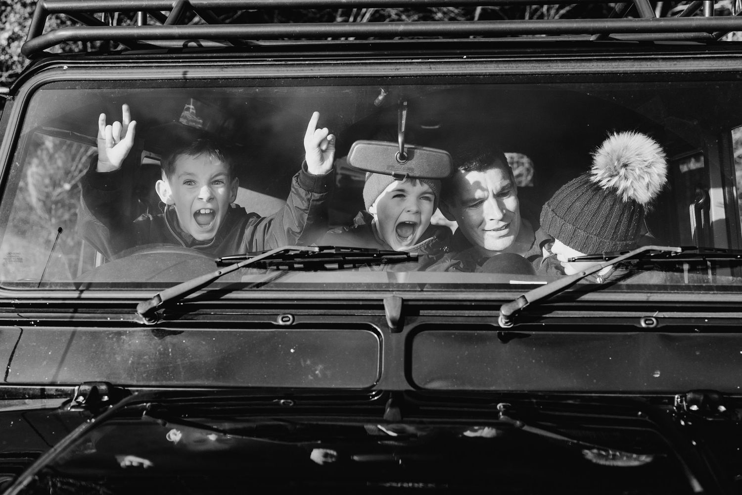 Black and white photo showing a group of people waving excitedly through the window of a vintage vehicle. family PHOTOGRAPHER Dublin