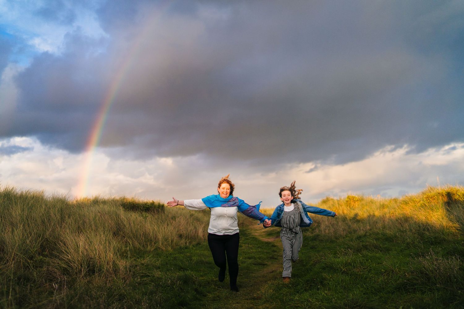 Two people hold hands while running through a grassy hilltop with a vibrant rainbow and dramatic clouds in the sky. family PHOTOGRAPHER Dubl