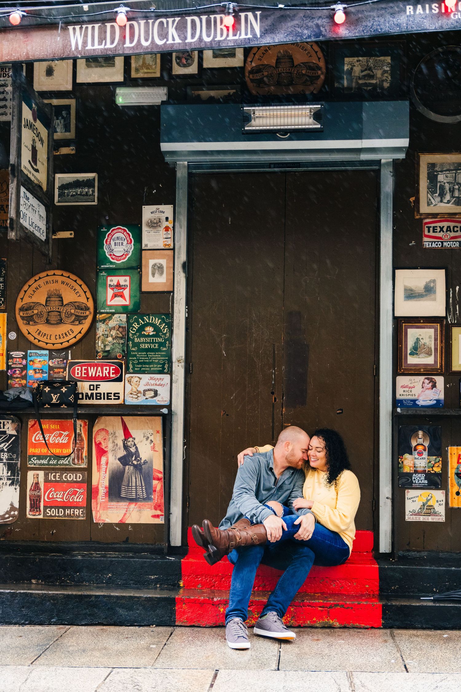 Couple sits on red steps outside Wild Duck Dublin pub surrounded by vintage signs and advertisements. PHOTOGRAPHER Dublin ireland