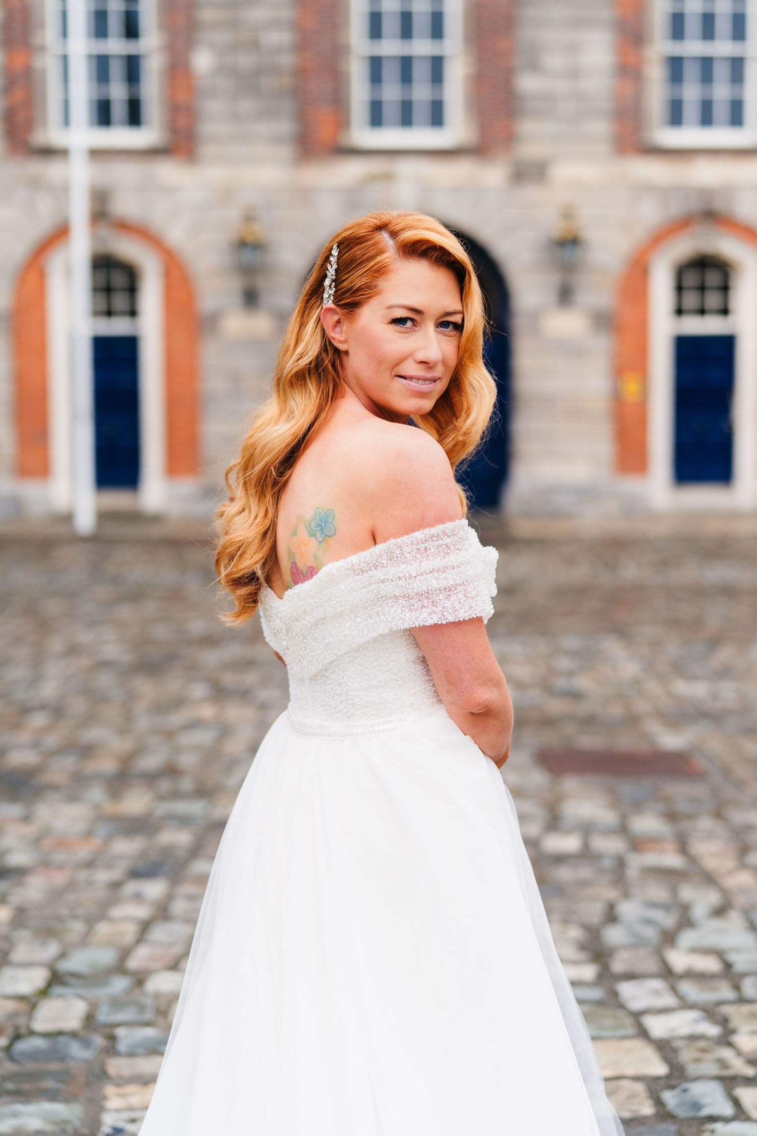 Bride in off-shoulder white gown poses on cobblestone courtyard in front of historic brick building.  city hall dublin wedding photos