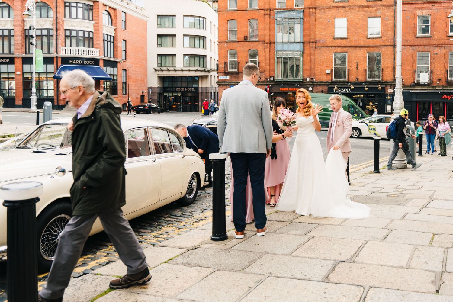 A wedding couple stands on a cobblestone street in Dublin with vintage cars and historic brick buildings in the background.  city hall dubli