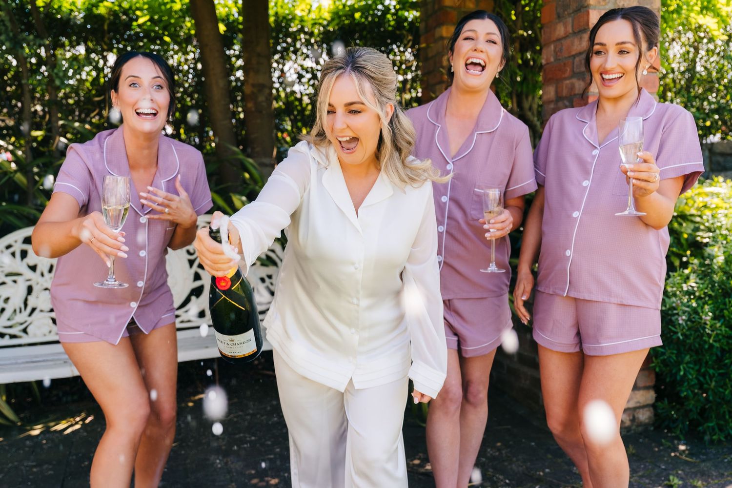 Wedding photographer Dublin Bride in white satin pajamas pops champagne with three bridesmaids in matching mauve pajama sets outdoors.