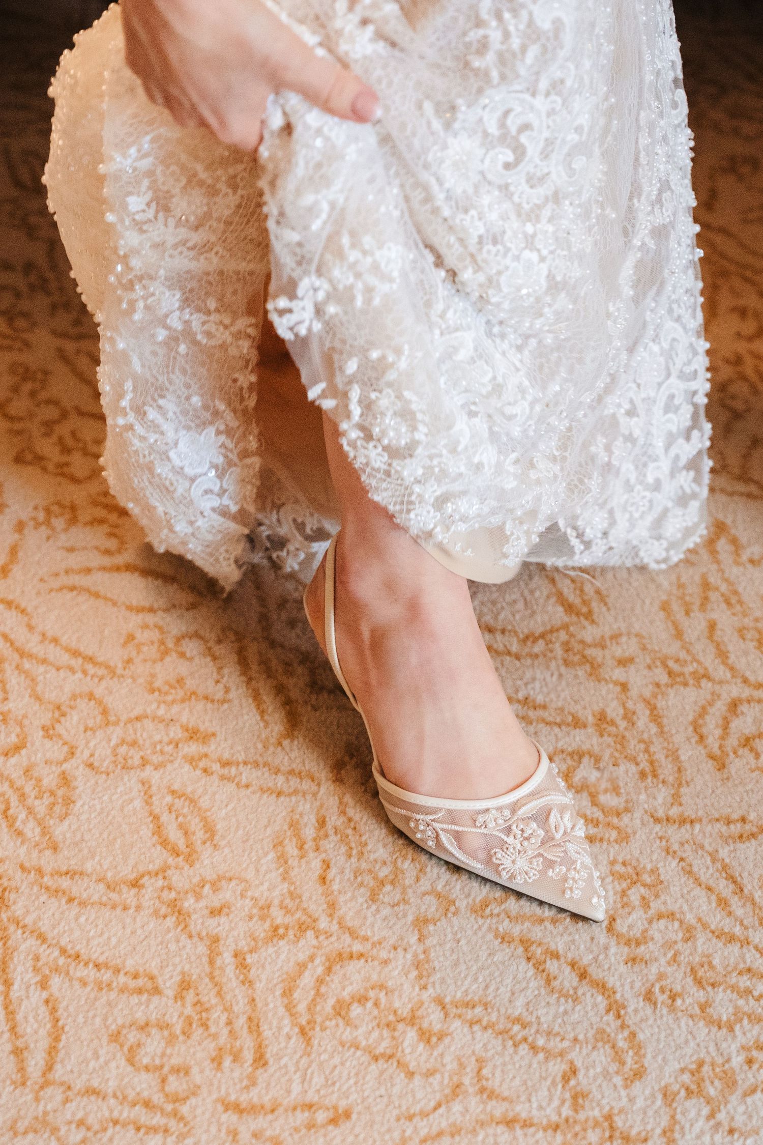 Elegant white lace wedding shoes with delicate details shown on a patterned carpet.