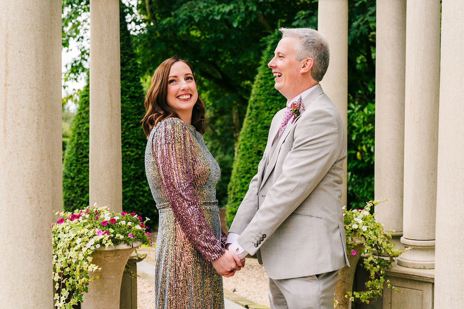 A couple shares a joyful moment while standing between white columns in a garden setting. Wedding Photographer Dublin
