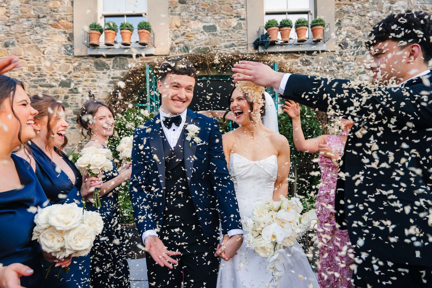 Newlyweds laugh joyfully as wedding guests shower them with confetti outside a stone building with flower boxes. Virginia Park Lodge Wedding