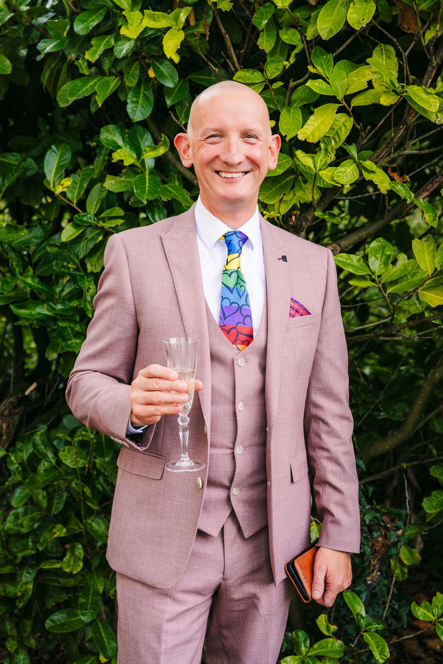 A person in a mauve three-piece suit with colorful tie holds a champagne glass against a leafy green background. LGBTQ Ashley Park Wedding P