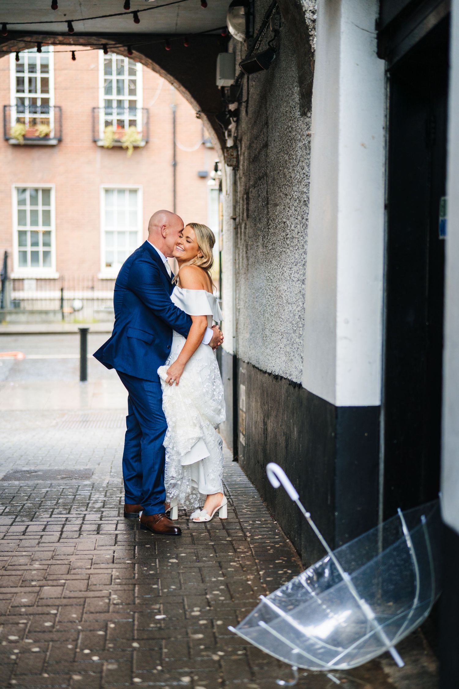 A couple shares an intimate moment under a walkway with an umbrella lying on the ground. The Dean Dublin Wedding Photographer