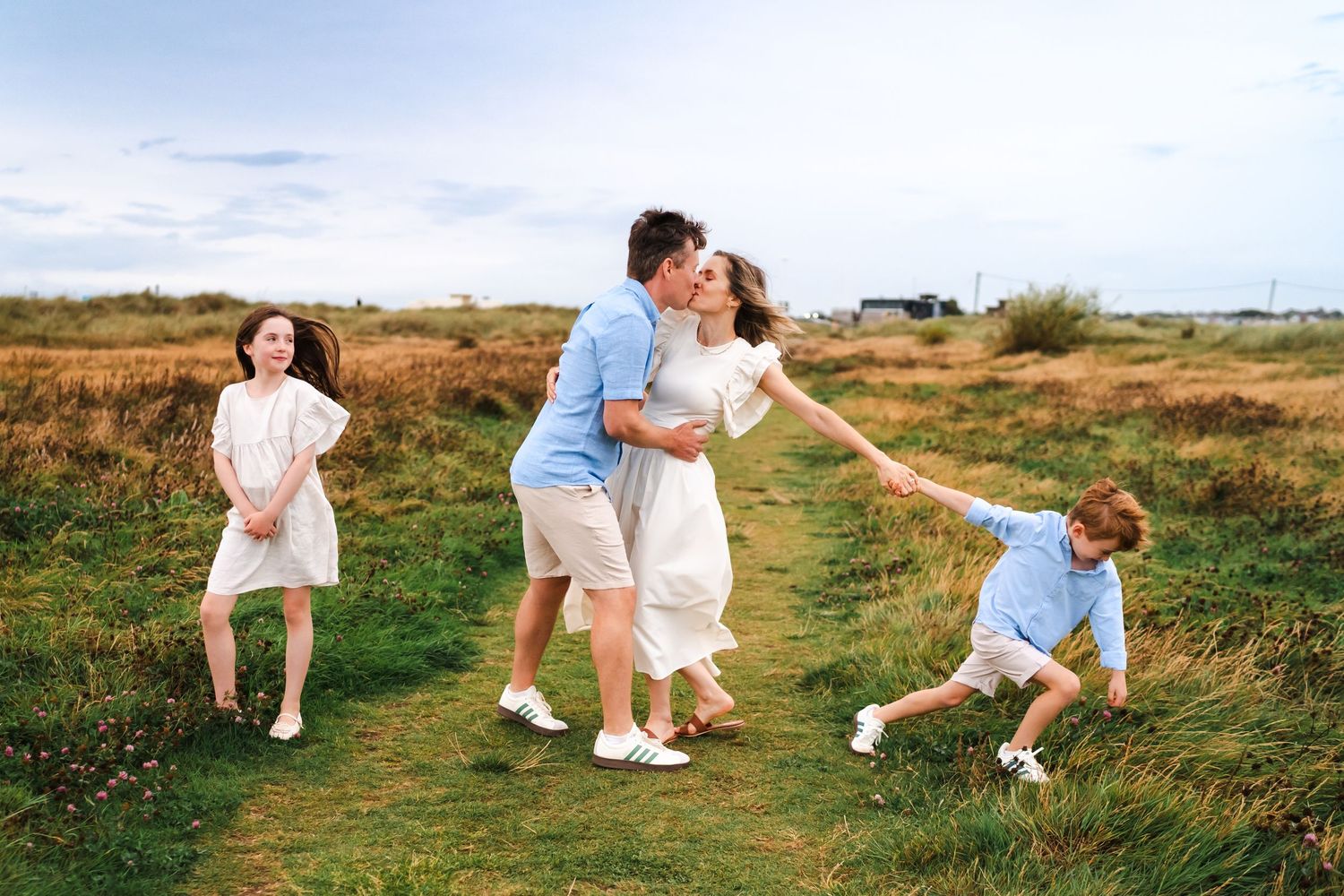 Two people in light summer clothing walk and play together in a sunny field with wildflowers and grass.  Family photographer Dublin Ireland