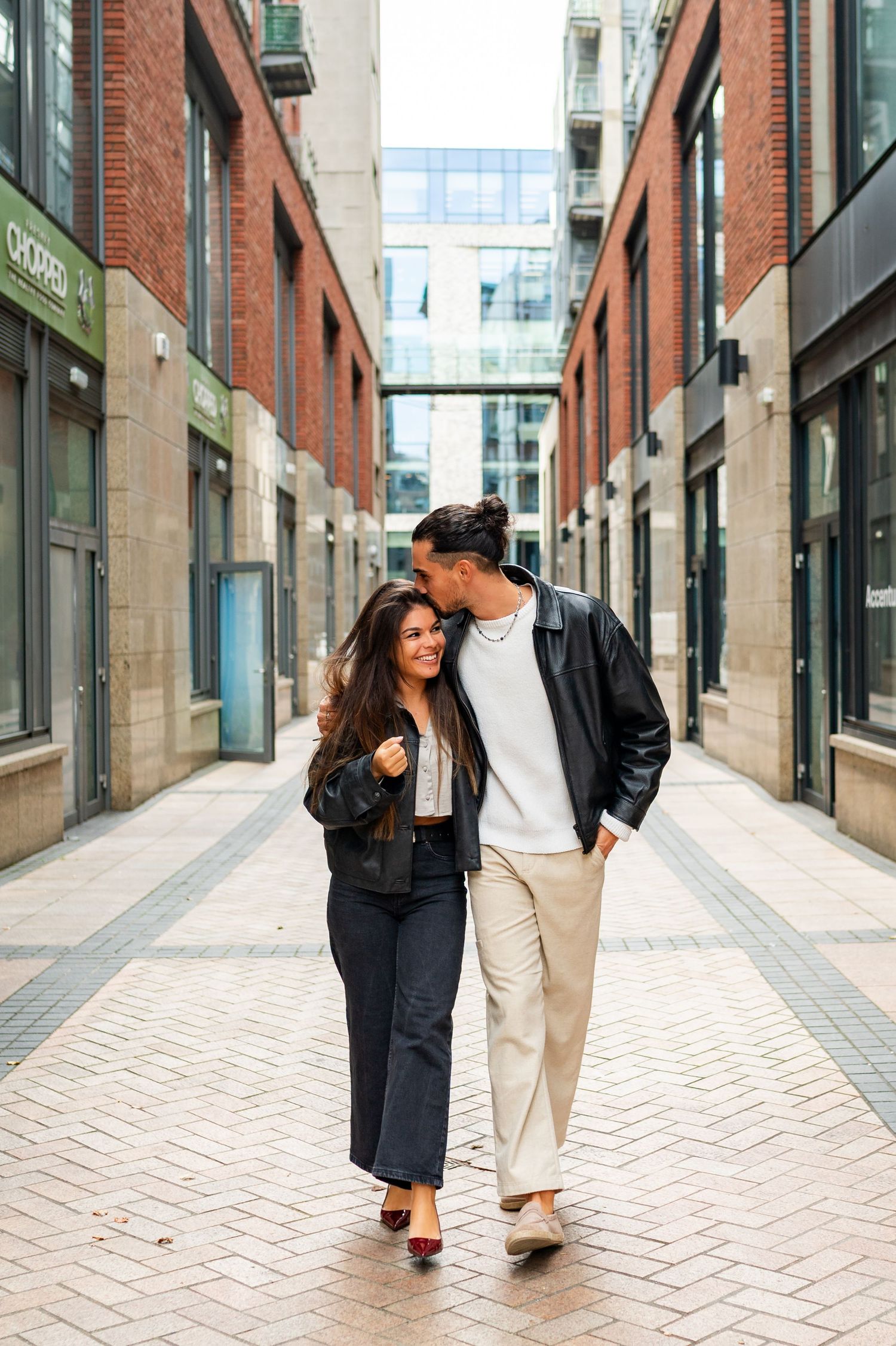A couple in contrasting outfits walks and poses playfully along a brick-lined urban street with storefronts. Couple's Photoshoot PHOTOGRAPHE