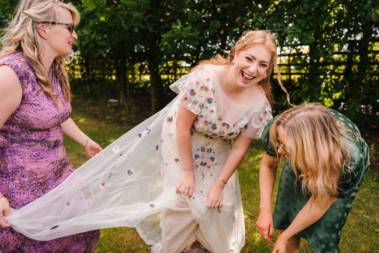 Guests help adjust a flowing wedding dress in a sunlit garden setting. . Village Barn Tyrrellspass wedding photographer Ireland