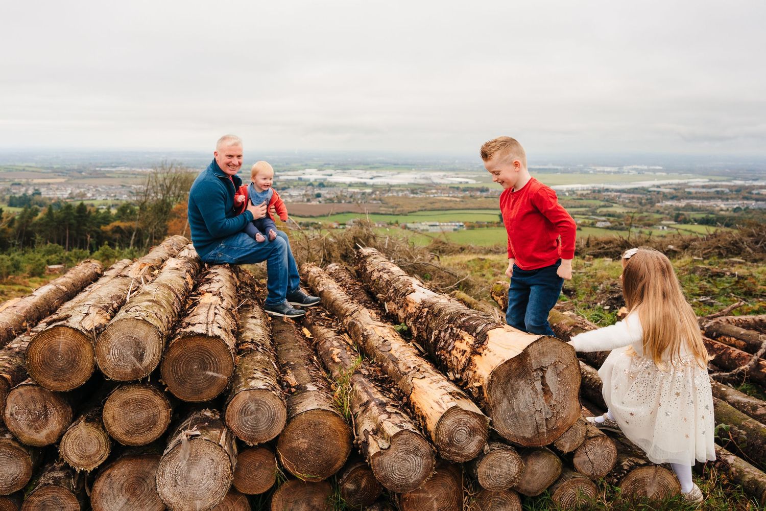 A happy couple poses on a large pile of stacked logs in a scenic mountainous forest location during sunset. Family Photographer Dublin Irela