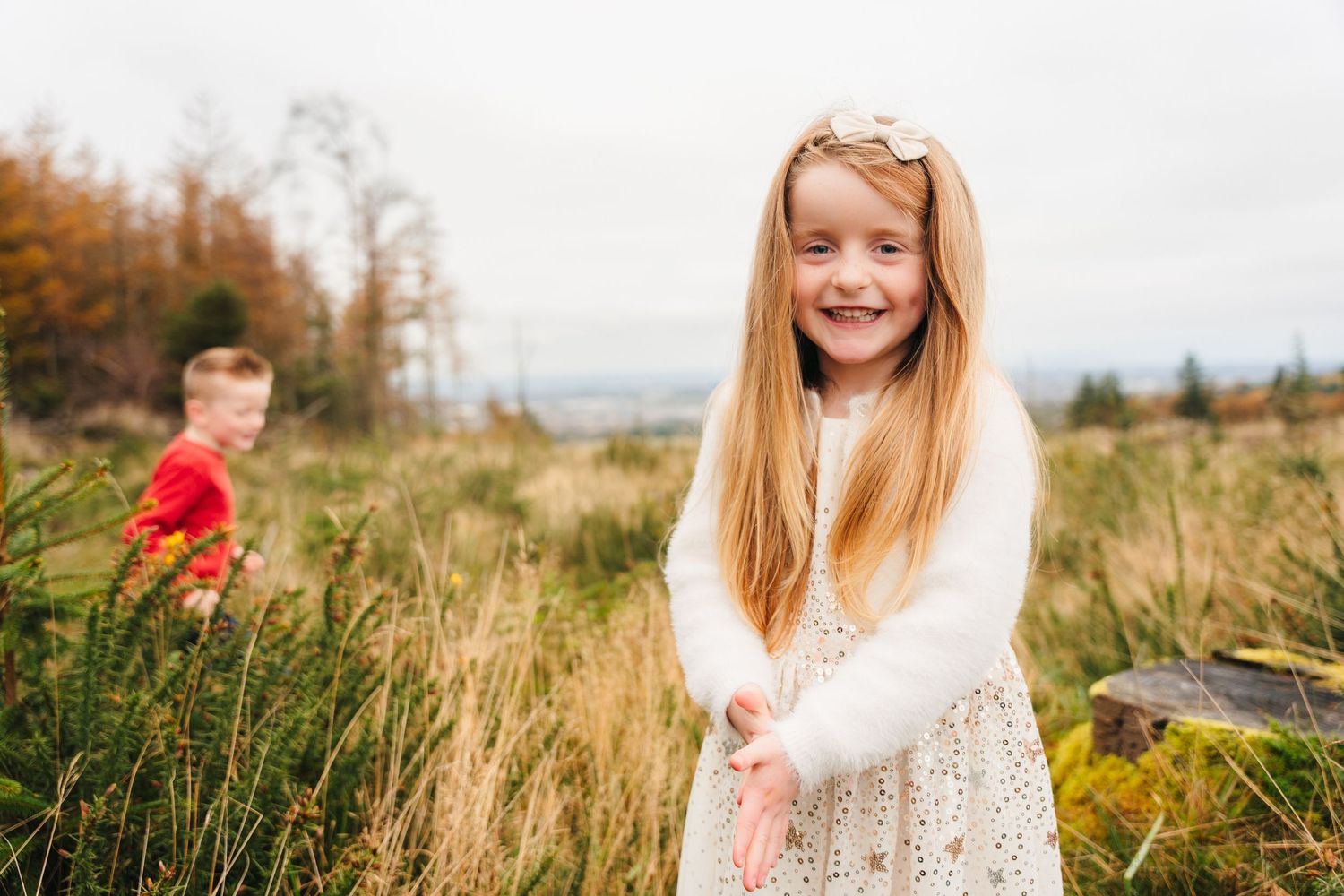 Family Photographer Dublin Ireland Young girl in white lace dress poses in tall grass during autumn with blurred figure in background.