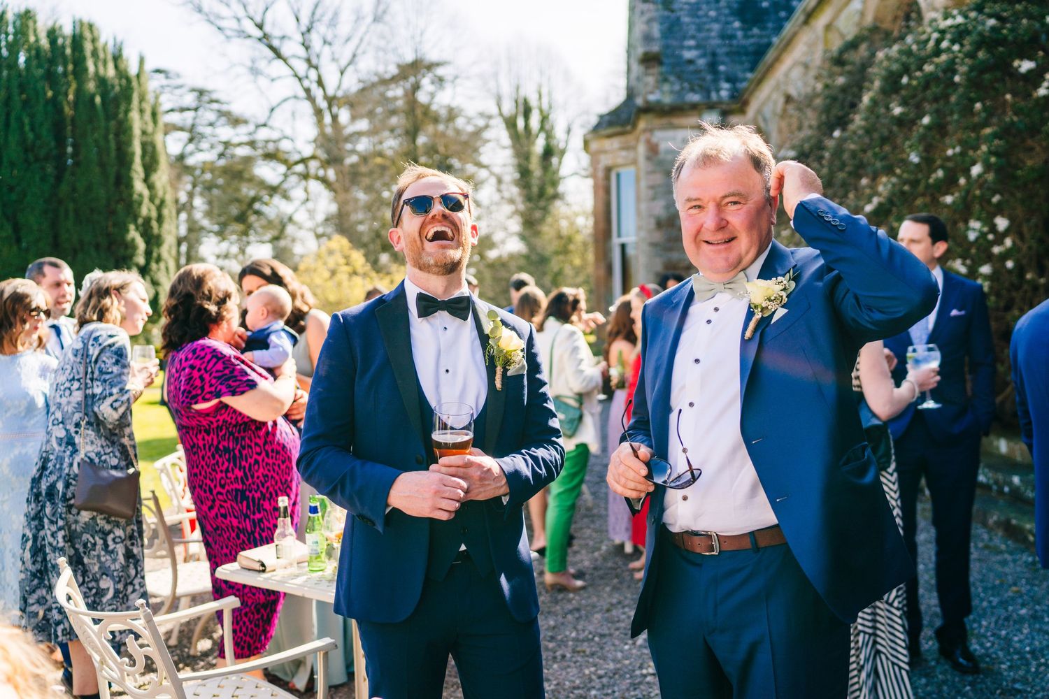 Castle Leslie WeddingTwo guests in formal blue suits laughing and celebrating at an outdoor wedding reception with other attendees visible.
