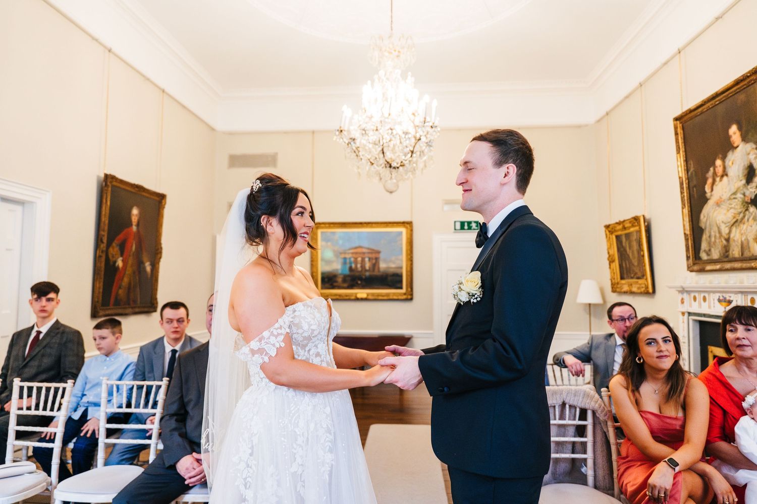 A wedding ceremony takes place in an elegant room with a crystal chandelier and white chiavari chairs. Wedding PHOTOGRAPHER Dublin