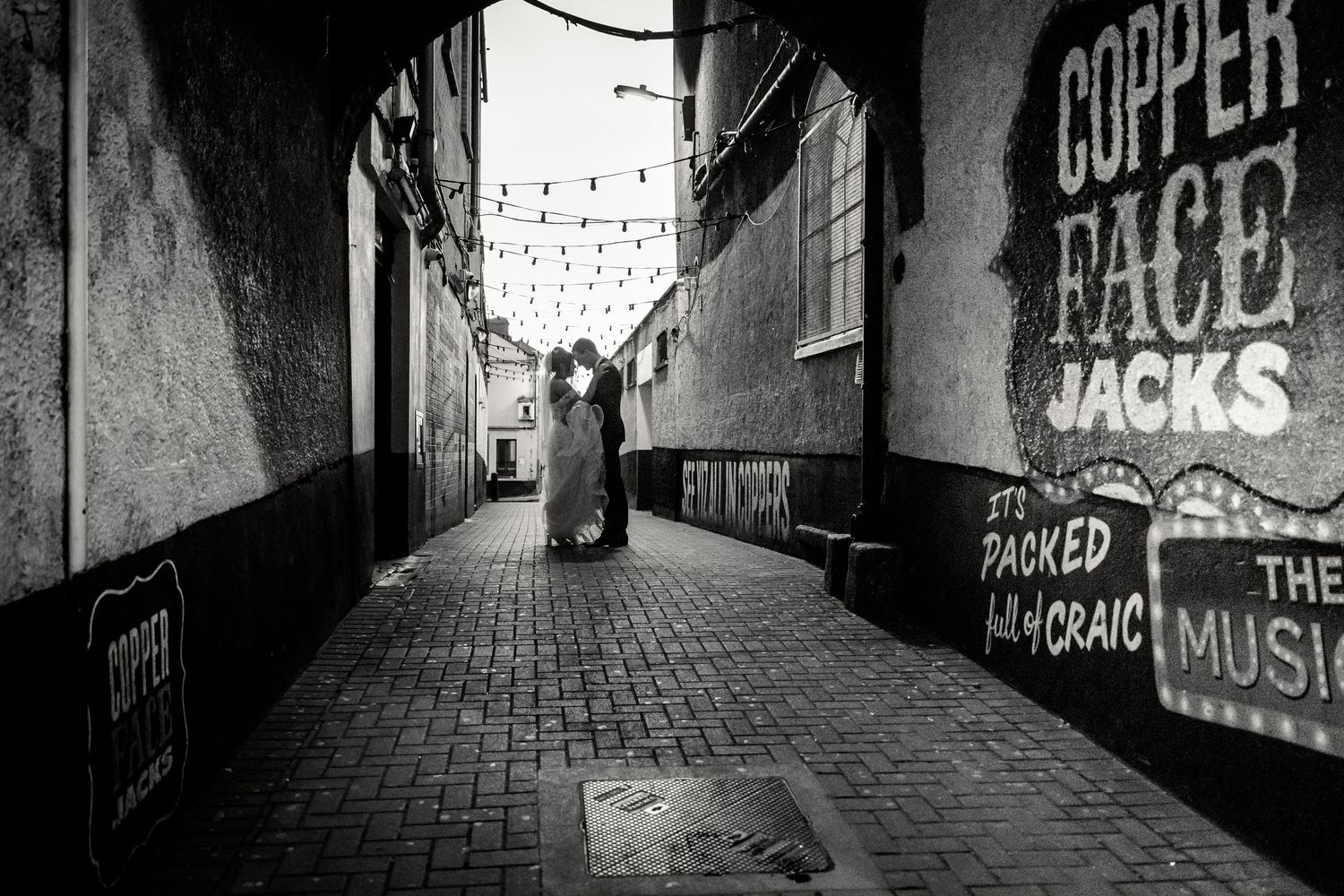 Black and white wedding photo in an urban alley with string lights overhead. Wedding PHOTOGRAPHER Dublin