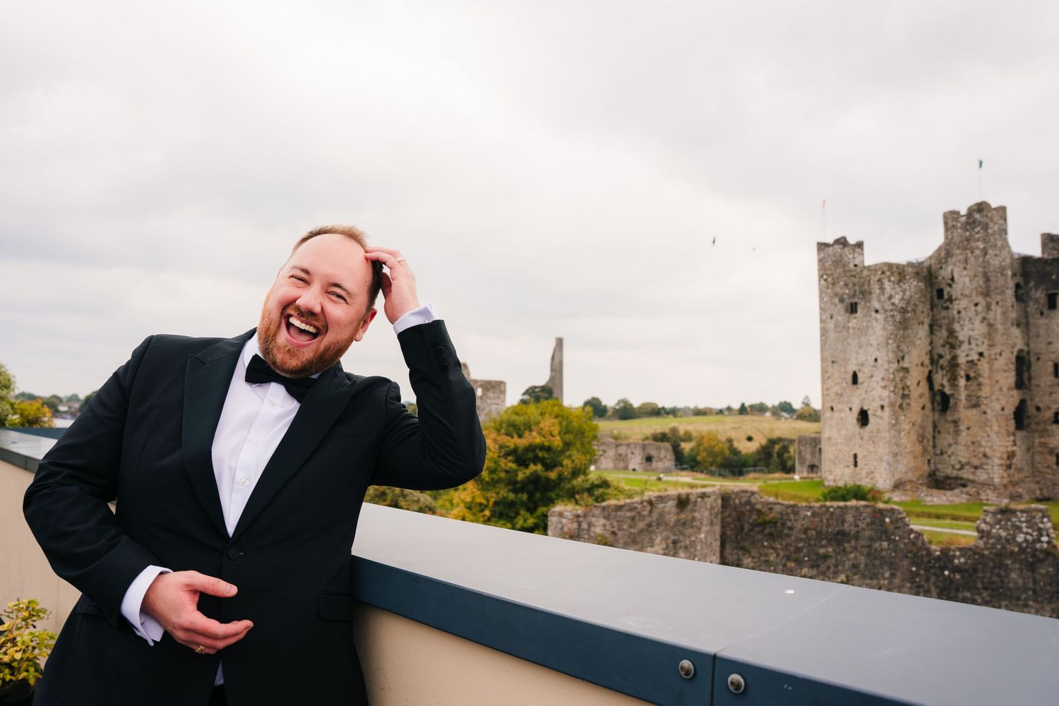 A person in a black suit laughs expressively while standing near a historic stone castle in Ireland. Wedding Photography Trim Castle Ireland