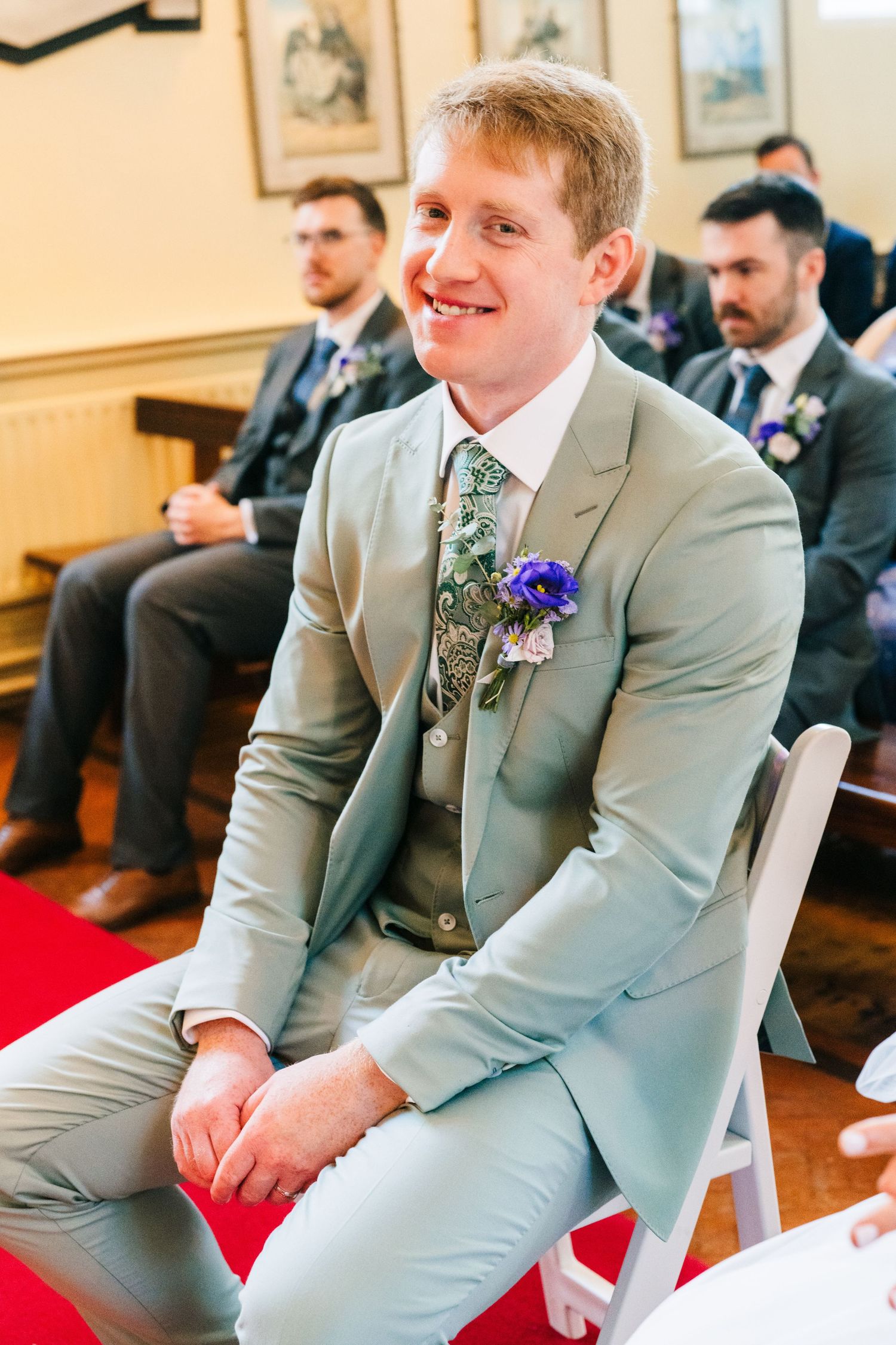 Millhouse slane wedding photograpLight grey suited wedding party member with floral tie and purple boutonniere sits smiling during ceremony.